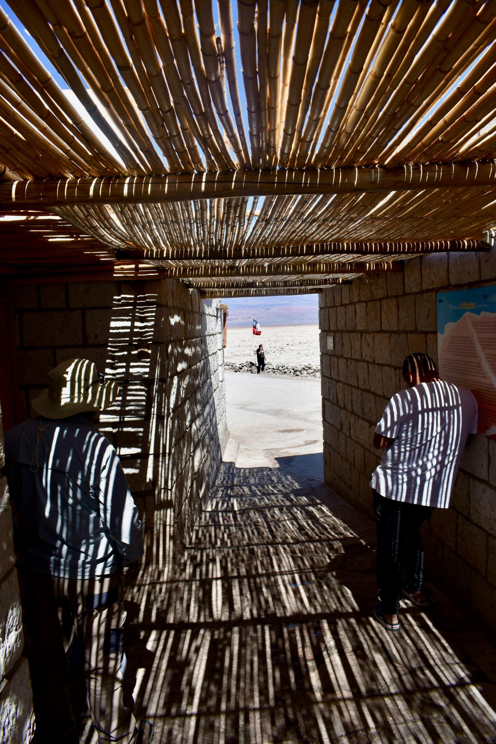 Shadows from bamboo roof create striped patterns in a tunnel leading outdoors.